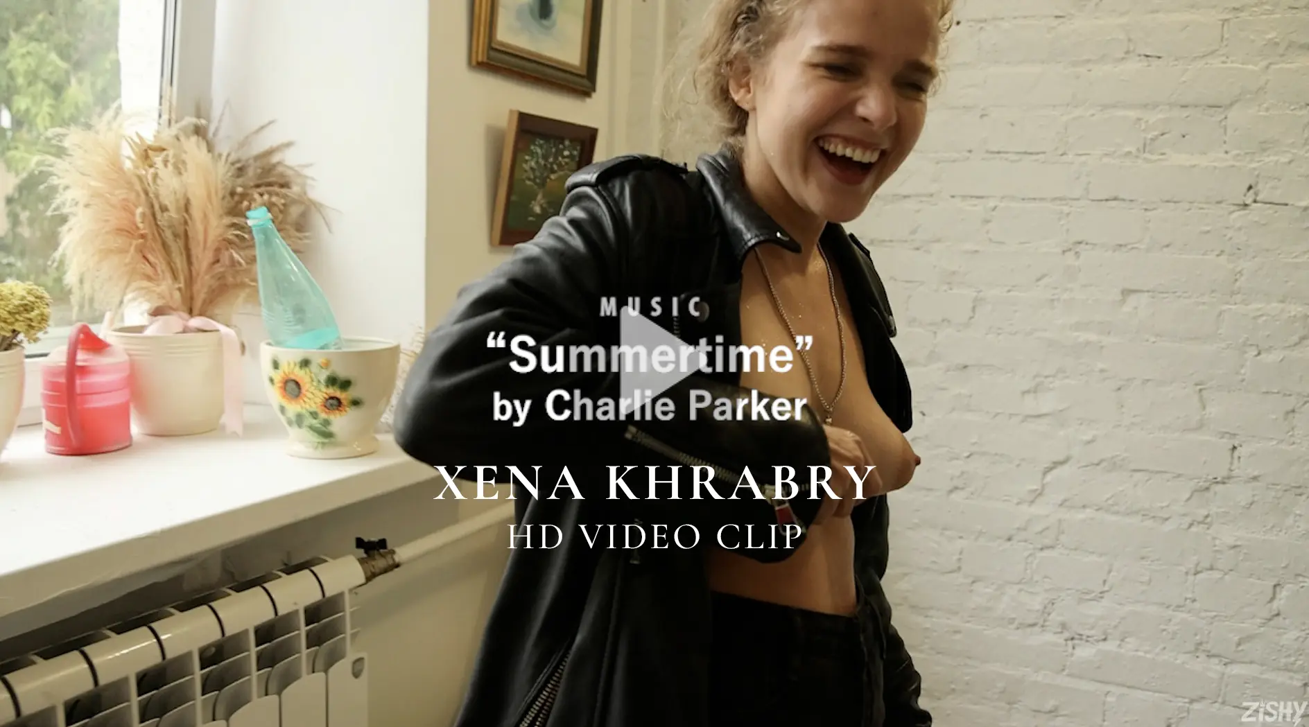 Xena smiling while opening her leather jacket and showing off her small breasts in the Zishy HD video preview